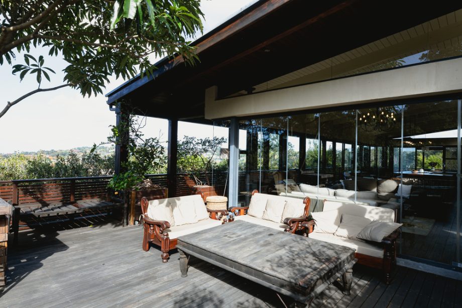 Elevated timber deck with white cushioned wooden furniture and floor-to-ceiling glass walls overlooking a tree-lined landscape.